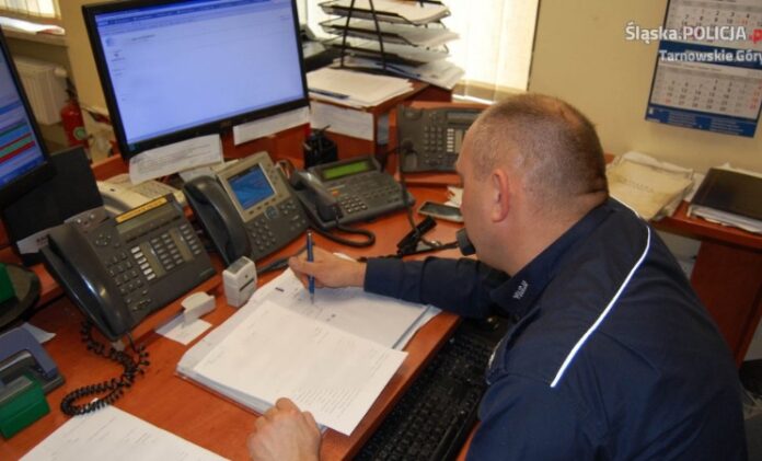 Na zdjęciu policjant siedzi przy biurku, patrzy w monitor komputera i rozmawia przez telefon.